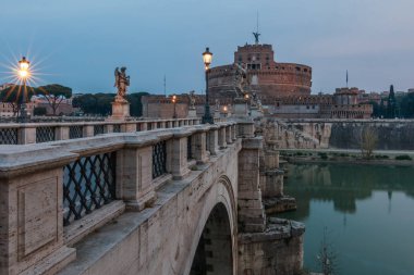 Sabah alacakaranlıkta Sant Angelo Kalesi ile Aurelius köprüsü. Taş köprünün yanal görünümü. Roma'da Tiber nehri üzerindeki köprüde ışık alan tarihi sokaklar