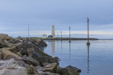 Deniz feneri ve arka planda ıssız evler. Suda yanal güç direkleri var. Gökyüzünde bulutlar ile Reykjavik yakınlarındaki Grotta adasının taş kıyı şeridi.