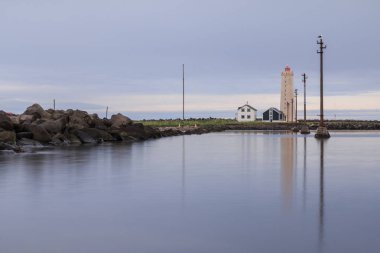 Deniz fenerine ve ıssız evlere suda güç direkleri ile Mavi gökyüzü. Reykjavik yakınlarındaki Grotta adasının taş kıyı şeridi bulutlu. Kuş barınağı olan yarımada