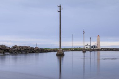 Reykjavik yakınlarındaki Grotta adasının taş kıyı şeridi bulutlu. Deniz fenerine ve ıssız evlere suda güç direkleri ile Mavi gökyüzü. Kuş barınağı olan yarımada