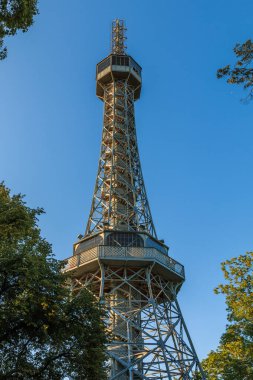 Prag 'da çelikten yapılmış eski Petrin radyo kulesi. Petrin Tepesi, Küçük Şehir Bölgesi' nde güneş ışığı ve mavi gökyüzünde gözlem güvertesi ve ön planda ağaçlarla birlikte.