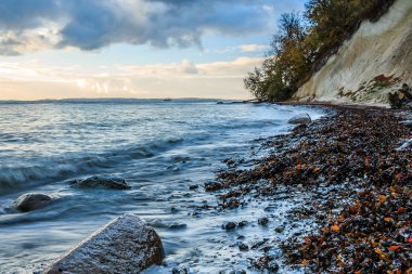 Baltık Denizi kıyısında. Ruegen adasındaki tebeşir kayalıkları. Yıkanmış deniz yosunlarıyla dolu taşlı bir sahil. Sonbahar havasında suda daha büyük taşlar. Dramatik bulutlarla gün doğumu 