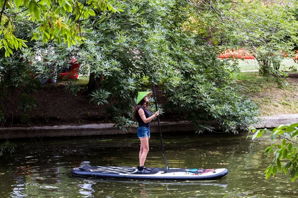 Moskova, Park Krasnaya Presnya, 05 Temmuz 2018: genç bir kız ile bir yandan çarklı ve hasır şapkalı bir gölet üzerinde sörf tahtası üzerinde duruyor.