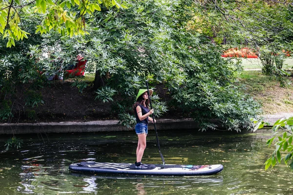 Moskova, Park Krasnaya Presnya, 05 Temmuz 2018: genç bir kız ile bir yandan çarklı ve hasır şapkalı bir gölet üzerinde sörf tahtası üzerinde duruyor.