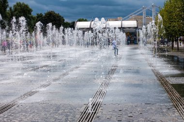 Küçük bir kız çeşmenin fıskiyeleri arasında koşar.