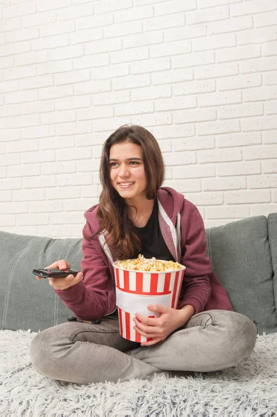 Teen girl sitting on couch with TV remote and eating popcorn — Stock Photo, Image