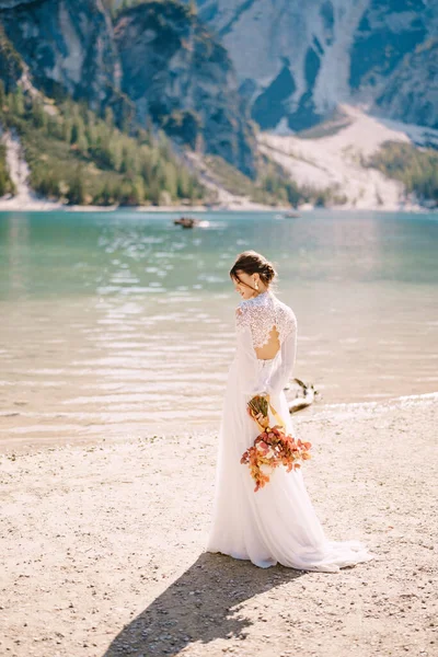 Bellissima sposa in abito bianco con maniche e pizzo, con un bouquet giallo autunnale di fiori secchi e rose peonate, sul Lago di Braies in Italia. Matrimonio di destinazione in Europa, sul lago di Braies . — Foto Stock