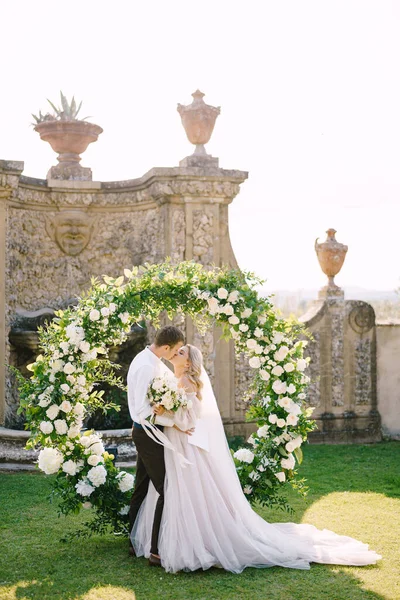 Wedding couple kisses. Wedding at an old winery villa in Tuscany, Italy. Round wedding arch decorated with white flowers and greenery in front of an ancient Italian architecture.