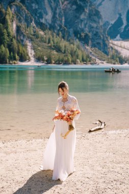 Kolları ve dantelli beyaz elbiseli güzel bir gelin, İtalya 'da Lago di Braies' de sarı bir sonbahar buketi ve şakayık gülleri ile. Düğün Avrupa 'da, Braies Gölü' nde..