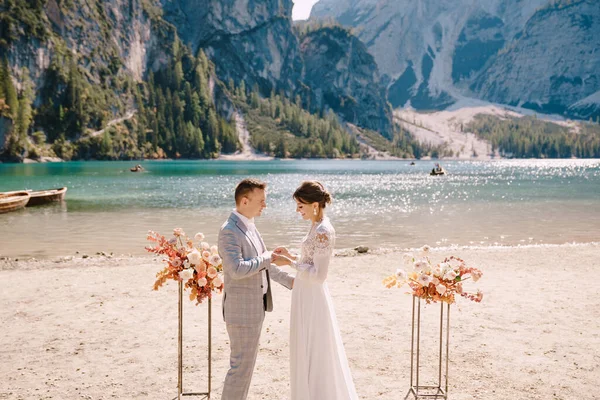 La sposa mette un anello sullo sposo in occasione della cerimonia, con un arco di colonne di fiori autunnali, sullo sfondo del Lago di Braies in Italia. Matrimonio di destinazione in Europa, sul lago di Braies . — Foto Stock
