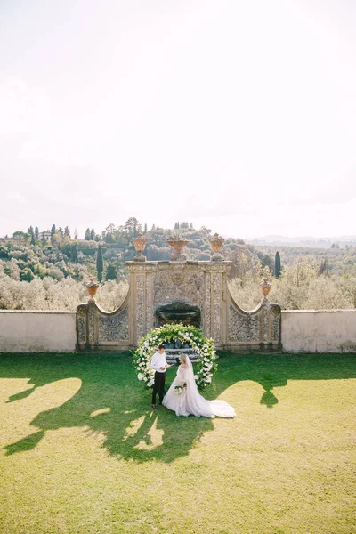 Boda en una antigua villa bodega en Toscana, Italia. Pareja de boda bajo un arco redondo de flores. El novio lee los votos de boda . —  Fotos de Stock