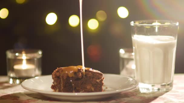 En fyrkantig bit choklad Brownie på en tallrik, hälls med gyllene saltad karamell. Mot bakgrund av brasan ljus, ett fullt glas mjölk och brinnande ljus. — Stockvideo