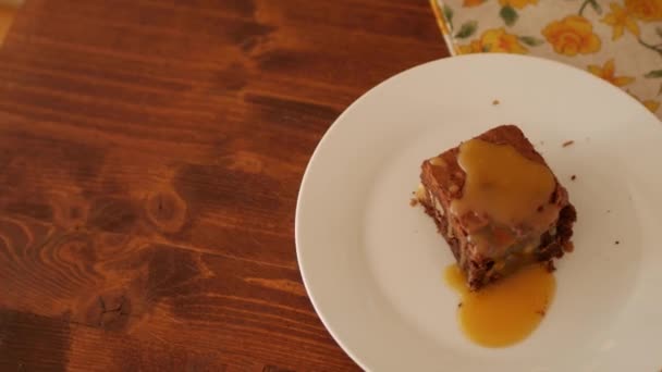 Un morceau carré de Brownie au chocolat sur une assiette, sur un fond en bois, une serviette en fleurs jaunes et une vieille cuillère melchior. Le caramel doré salé s'égoutte lentement . — Video