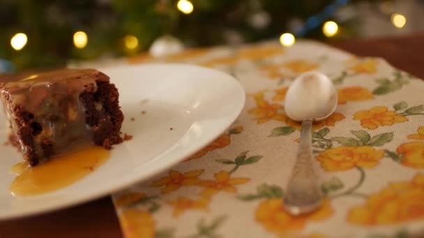 Een vierkant stukje Chocolade Brownie op een bord, op een houten achtergrond, een servet in gele bloemen en een oude melchior lepel. Goudgezouten karamel lekt langzaam af. — Stockvideo