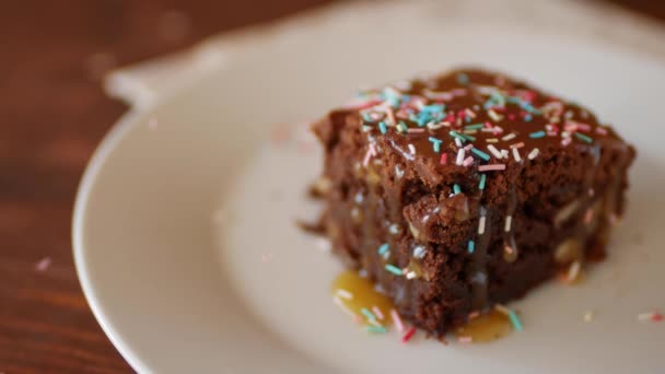 Um pedaço quadrado de chocolate Brownie em um prato polvilhado com pó de pastelaria multi-colorido . — Vídeo de Stock