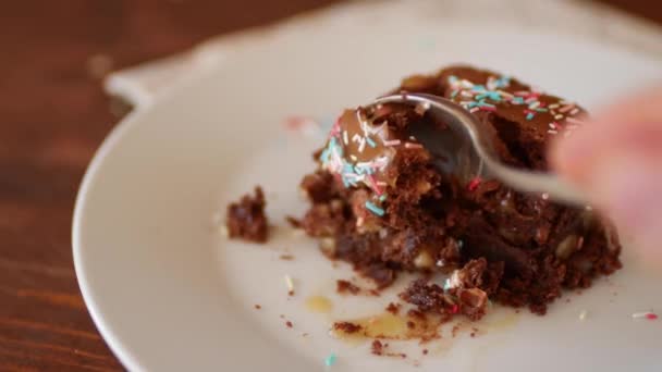 Een vierkant stukje chocolade Brownie op een bord, bestrooid met veelkleurig suikerpoeder, eet een meisje met een lepel. — Stockvideo