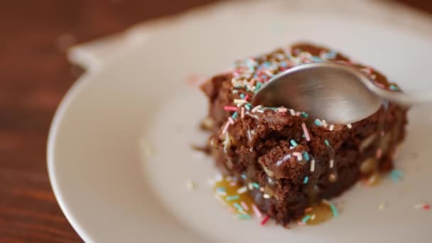 Een vierkant stukje chocolade Brownie op een bord, bestrooid met veelkleurig suikerpoeder, eet een meisje met een lepel. — Stockvideo