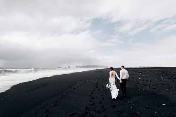 Ett bröllopspar går längs den svarta stranden i Vic. Sandstrand med svart sand på Atlantens stränder. Brud och brudgum håller hand. Destination Island bröllop. — Stockfoto