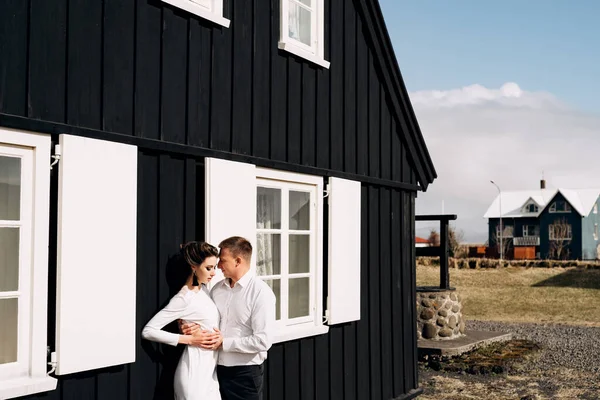 Svatba na Islandu. Svatební dvojice u černého dřevěného domu s bílými okny a okenicemi. Ženich objímá nevěstu. — Stock fotografie