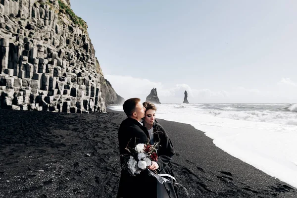 Un couple de mariés se promène le long de la plage de sable noir de Vik, près du basalte, sous la forme de piliers. Les mariés s'étreignent sur le rivage. Destination Mariage Islande . — Photo