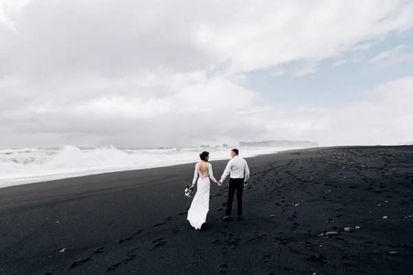 Un couple de mariés marche le long de la plage noire de Vic. Plage de sable avec sable noir sur les rives de l'océan Atlantique. Mariée et marié tenant la main. Destination Mariage Islande . — Photo