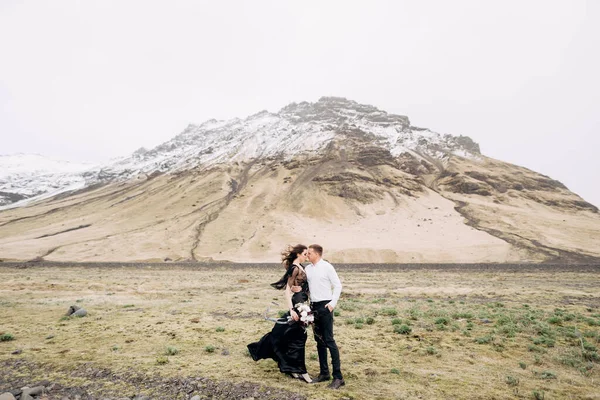 Couple de mariage sur fond de montagnes enneigées. La mariée dans une robe noire et marié sont câlins dans un champ de mousse et d'herbe jaune. Destination Mariage Islande . — Photo