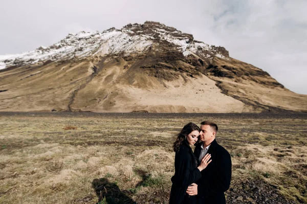 Mariage Destination Islande. Couple de mariage sur fond de montagnes enneigées. Les mariés en manteaux noirs s'étreignent dans un champ de mousse et d'herbe jaune . — Photo