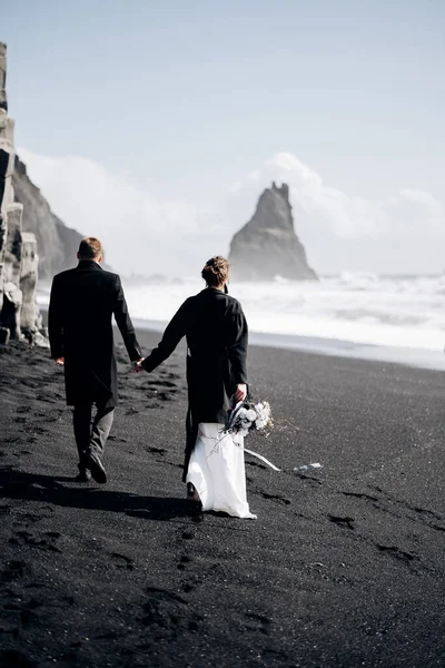 Mariage Destination Islande. Un couple de mariés se promène le long de la plage de sable noir de Vik, près du basalte, sous la forme de piliers. Les mariés se tiennent la main et marchent le long du rivage . — Photo