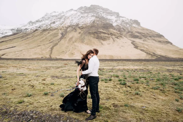 Couple de mariage sur fond de montagnes enneigées. La mariée dans une robe noire et marié sont câlins dans un champ de mousse et d'herbe jaune. Destination Mariage Islande . — Photo