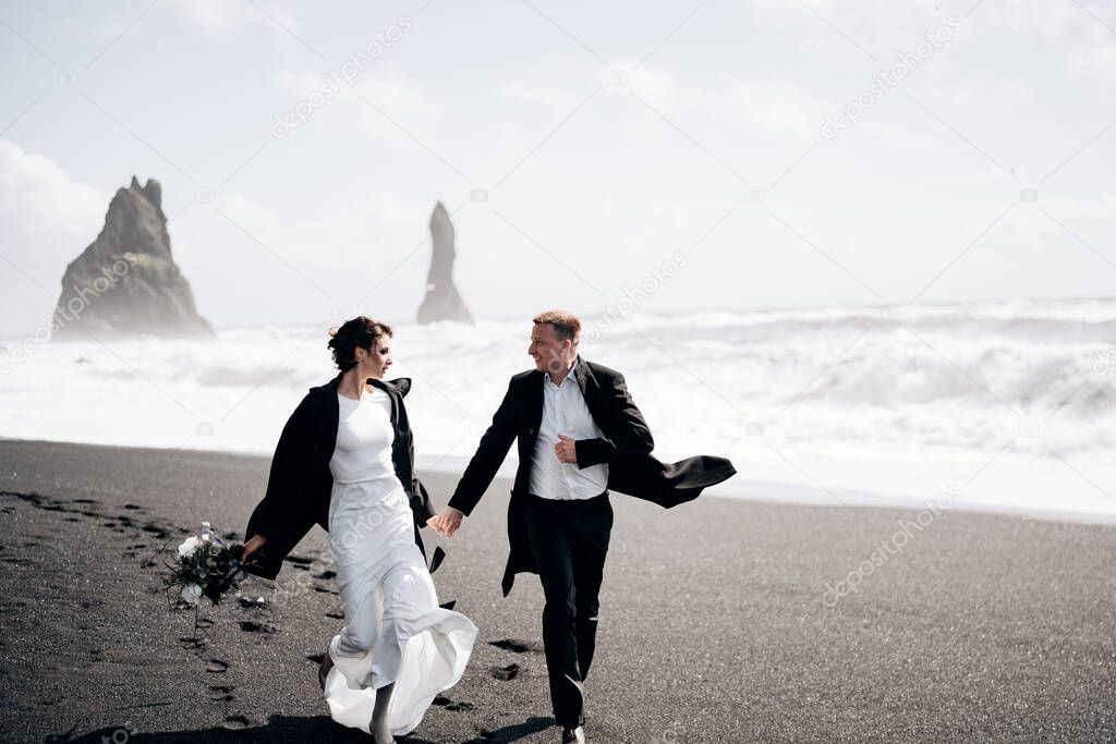 Destination Iceland wedding. The wedding couple runs along the sandy black beach of Vik, near the basalt rock, in the form of pillars. Holding hands, running along the shore against backdrop of waves