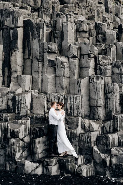 Un couple de mariés se tient sur un mur de piliers de pierre. Les mariés s'étreignent sur le basalte Kekurs, sur la plage de sable noir de Vik. Destination Mariage Islande . — Photo