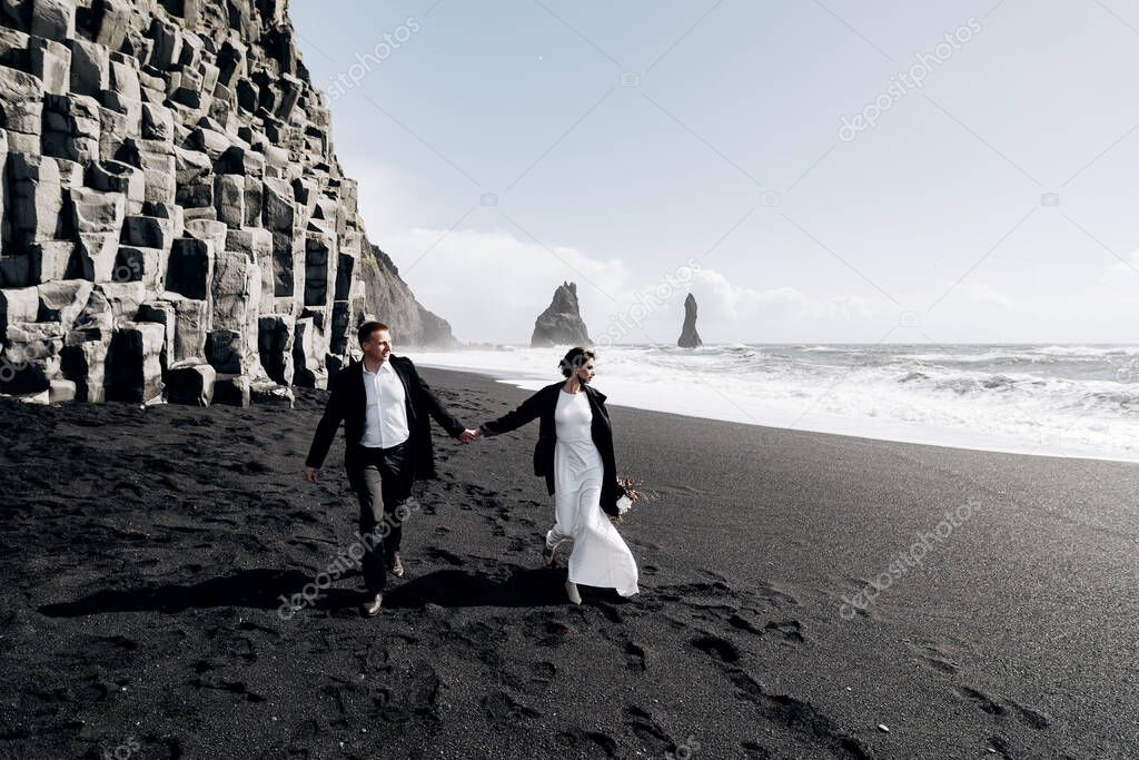 Destination Iceland wedding. A wedding couple walks along the sandy black beach of Vik, near the basalt rock, in the form of pillars. The bride and groom hold hands and walk along the shore.
