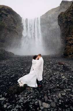 Skogafoss Şelalesi yakınlarında bir düğün çifti. Gelin ve damat yün bir battaniyeyle örtülmüş, orada sarılıyorlar. Kar yağıyor, kar yağıyor..