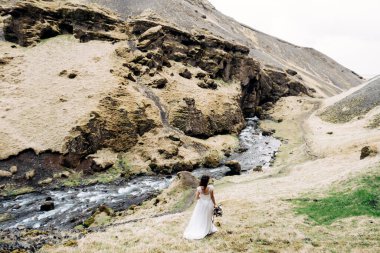 Beyaz elbiseli bir gelin ve elinde bir buket çiçek güney İzlanda 'daki Kvernufoss şelalesinin yanındaki bir dağ nehrinin kıyısında yürüyor..