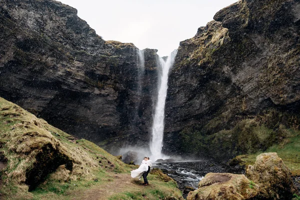 Ο γαμπρός κυκλώνει τη νύφη στην αγκαλιά του κοντά στον καταρράκτη, χιονίζει. Προορισμός Ισλανδία γάμος, κοντά Kvernufoss καταρράκτη. — Φωτογραφία Αρχείου
