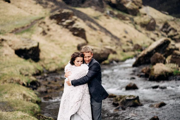 Um casal de casamentos fica na margem de um rio de montanha sob um cobertor de lã. O noivo abraça a noiva, tentando aquecê-la. A neve cai. Casamento destino Islândia, perto de cachoeira Kvernufoss . — Fotografia de Stock