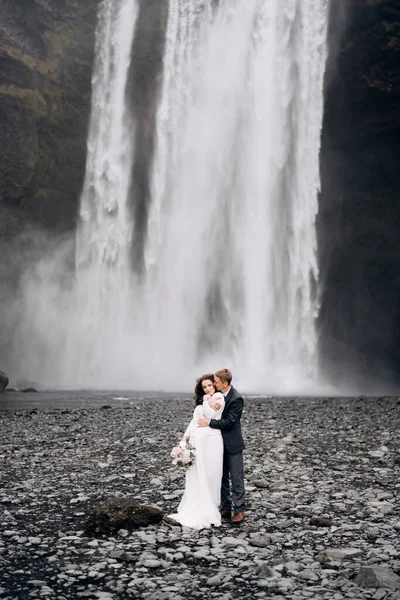 De bruidegom knuffelt de bruid van achteren. Bruidspaar bij de Skogafoss waterval. Reisdoel IJsland bruiloft. — Stockfoto