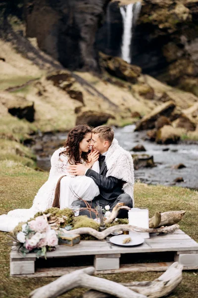 Mariage Destination Islande, près de la cascade de Kvernufoss. Un couple de mariés est assis sur les rives d'une rivière de montagne, à une table pour un dîner de mariage, faite d'une palette en bois peinte avec de la mousse et des chicots — Photo