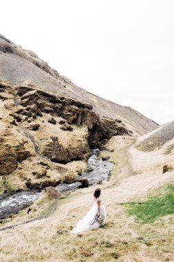 Beyaz elbiseli bir gelin ve elinde bir buket çiçek güney İzlanda 'daki Kvernufoss şelalesinin yanındaki bir dağ nehrinin kıyısında yürüyor..