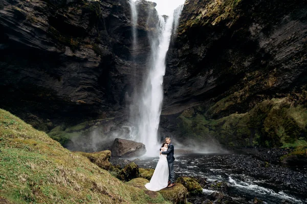 Tujuan pernikahan Islandia, dekat air terjun Kvernufoss. Pasangan pengantin berdiri di dekat air terjun. Pengantin pria memeluk pengantin wanita. — Stok Foto