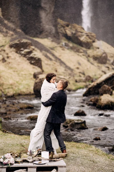 Reisdoel IJsland bruiloft, nabij Kvernufoss waterval. Bruidspaar aan de oever van een bergrivier. De bruidegom draagt de bruid in een wollen deken in zijn handen.. — Stockfoto