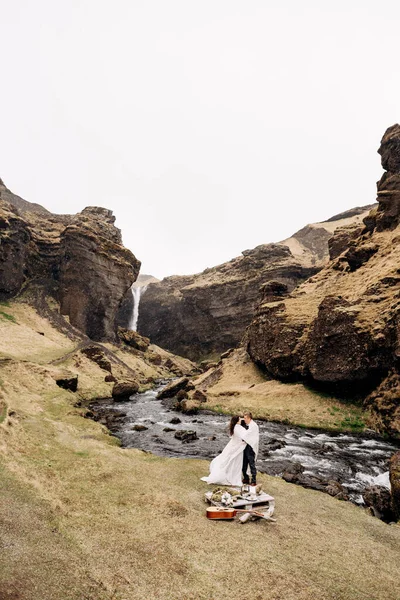 Reisdoel IJsland bruiloft, nabij Kvernufoss waterval. Een bruidspaar staat onder een ruit bij een bergrivier. De bruidegom knuffelt bruid. Ze bouwden een geïmproviseerde bruiloft tafel met decor en gitaar — Stockfoto