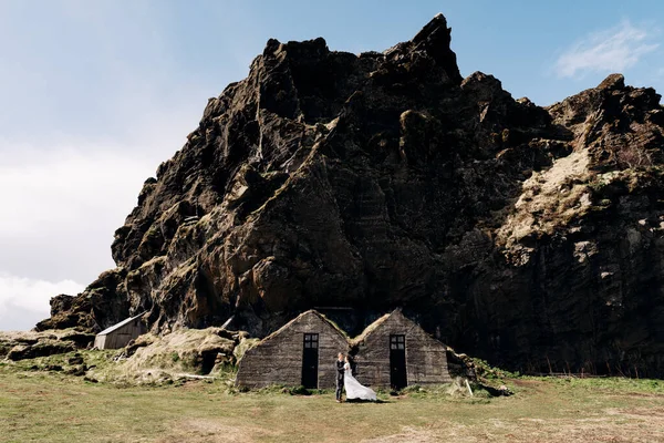 Bruidspaar poserend bij de oude huizen, bedekt met mos, op de achtergrond van een rotsachtige berg. Reisdoel IJsland bruiloft. — Stockfoto