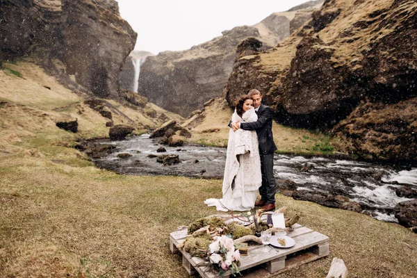 Casamento destino Islândia, perto da cachoeira Kvernufoss. Um casal de casamentos fica sob uma planície perto de um rio de montanha. O noivo abraça a noiva. Eles construíram uma mesa de casamento improvisada com decoração e guitarra — Fotografia de Stock