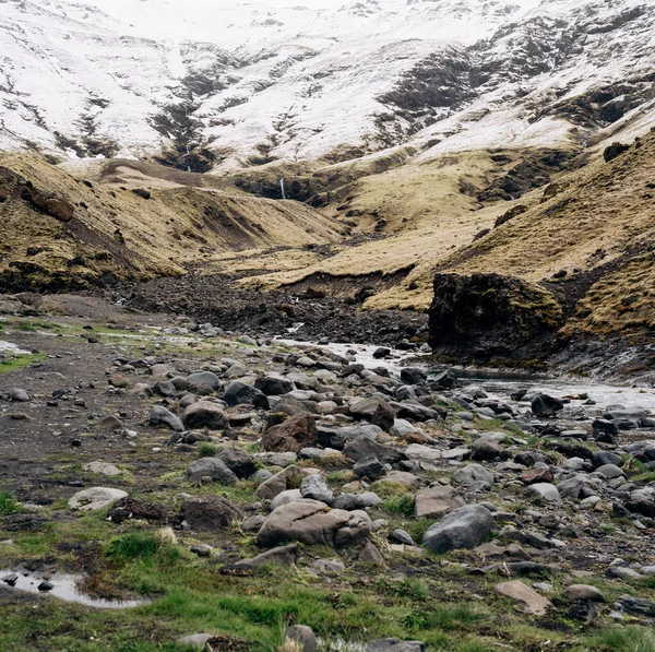 Malá mělká horská říčka, teče ze zasněžených hor do rokle, na kaskádě vodopádů. Sněhem pokrytý vrchol hory a chodidlo ve žluté suché trávě. — Stock fotografie