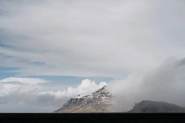 Um amplo campo de grama amarela seca e uma montanha à distância com um pico coberto de neve, contra um céu azul com nuvens brancas na Islândia . — Fotografia de Stock
