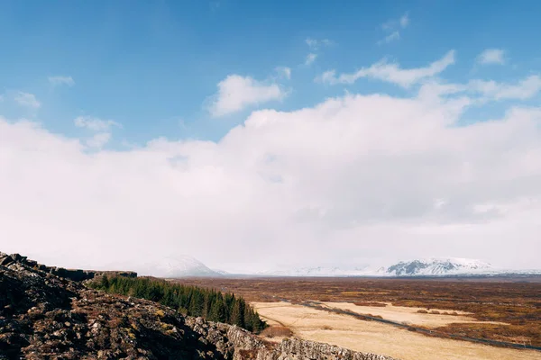 Utsikt från Silfra Fault, i Tingvedlir Valley på Island, till de stora vidderna av fält med gult gräs, barrskog, snötäckta bergstoppar och blå himmel med vita moln. — Stockfoto