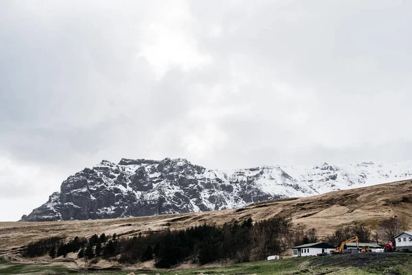 Білі будинки і будівельний екскаватор біля лісу на пагорбі, на тлі скелястої засніженої гори в Ісландії.. — стокове фото