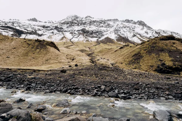 Hegyi folyó a hegy lábánál, hófödte csúccsal. Sárga száraz fű a hegyekben májusban Izlandon. — Stock Fotó