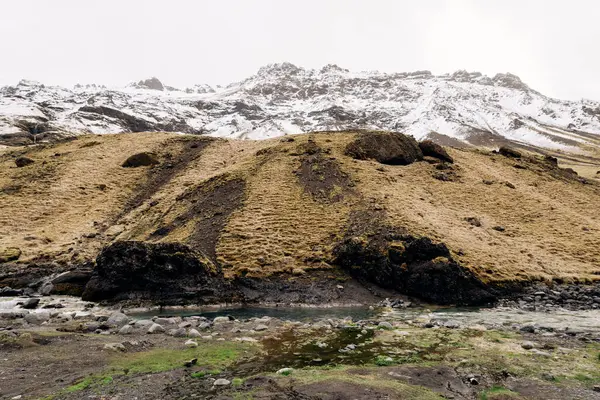 Rivière de montagne au pied de la montagne avec un pic enneigé. Herbe sèche jaune sur les montagnes en mai en Islande . — Photo